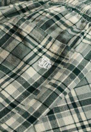 Tkanina flanelowa w kratę w odcieniach zieleni i beżu o teksturowanym wykończeniu. W rogu znajduje się białe haftowane logo.