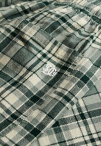 Tissu en flanelle à carreaux aux tons verts et beige avec une finition texturée. Présente un logo brodé blanc dans un coin.