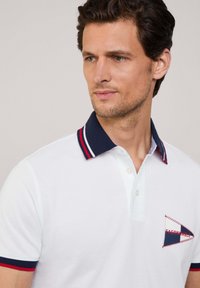 Camisa polo blanca con cuello azul marino que presenta rayas rojas y blancas. Tiene un logo trianguloso bordado en azul y rojo en el pecho.