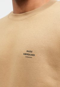 Közelkép egy bézs ing dekoltázsáról és melléről, amelyen kis fekete felirat szerepel: "Mads Nørgaard Copenhagen."