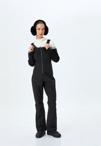 Zwarte skioverall met verstelbare banden, ritssluiting aan de voorkant en taps toelopende benen. Gecombineerd met een wit en zwart hoodie-top en zwarte oorkappen.