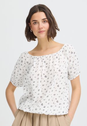 Frau mit kurzen braunen Haaren, die silberne Creolen trägt und eine weiße, kurzärmelige Bluse mit Blumenmuster sowie beige Hosen, Hände in den Taschen.