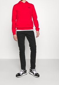 Roter Kapuzenpullover mit Zugband und schwarzem Logo, getragen über einem weißen Shirt; kombiniert mit schwarzen Slim-Fit-Jeans und schwarz-weißen Sneakers.