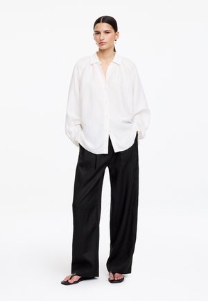 Kobieta stoi w luźnej białej koszuli na guziki, czarnych szerokich spodniach i czarnych japonkach na tle jednolitego białego tła.
