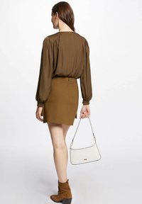 Blusa marrone a maniche lunghe con spalle plissettate, abbinata a una mini gonna marrone. La modella tiene in mano una borsa a tracolla bianca con texture. Stivali alla caviglia in suede marrone.