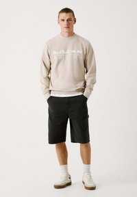 Sudadera beige con letras en blanco "Red Bull Racing", combinada con pantalones cortos oscuros. Calcetines y zapatillas blancas completan el conjunto. Look casual y deportivo.