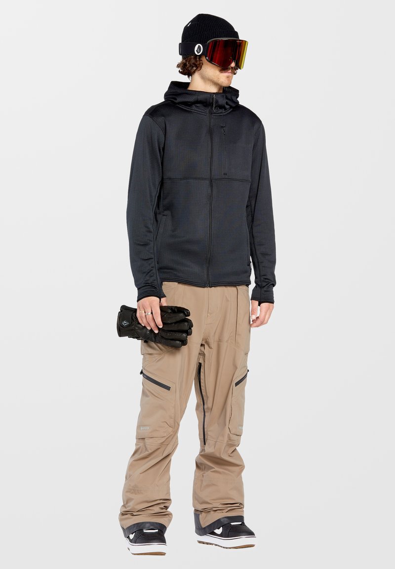 Schwarze Zip-Hoodie mit strukturiertem Muster, beiges Schneehosen mit Reißverschluss-Taschen, schwarze Handschuhe und Skibrille mit verspiegeltem Glas.