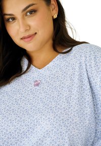 Jasnoniebieska koszula w kwiatowy wzór z dekoltem w szpic i krótkimi rękawami, ozdobiona małą różową kokardką na wysokości klatki piersiowej. Gładka faktura tkaniny.