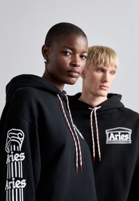 Zwarte hoodies met een witte "Aries" logo en grafisch ontwerp op de mouw, met gestructureerde koordjes in rood en wit. Casual stijl, ontspannen pasvorm.