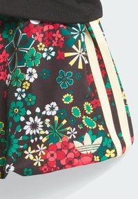 Μαύρο ύφασμα με λουλουδένιο μοτίβο που περιλαμβάνει κόκκινα, πράσινα και κίτρινα λουλούδια· διαθέτει πλευρικές ρίγες και λογότυπο με τρία φύλλα.