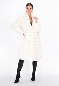 Witte faux fur jas met een pluche, geribde structuur en een brede kraag. Heeft een ontspannen pasvorm en lange mouwen, gecombineerd met strakke, zwarte leggings.
