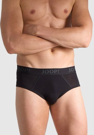 Mann mit verschränkten Armen, trägt schwarze Shorts mit "JOOP!"-Logo auf dem Bund vor einfarbigem Hintergrund.