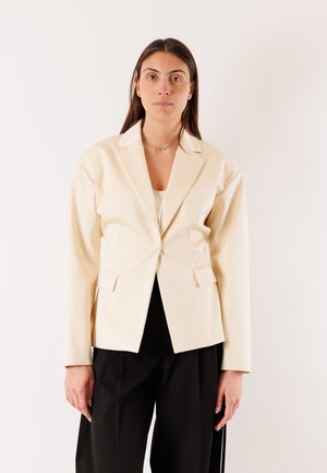 Donna con capelli lunghi castani che indossa un blazer color crema e pantaloni neri, in piedi davanti a uno sfondo bianco semplice, rivolta in avanti.