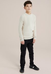 Sweater din tricot bej deschis, cu manșete ribionate, asortat cu pantaloni cargo negri cu buzunare și adidași negri cu accente albe.