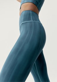 Detailn í záběr na osobu v vysokých modrozelených legínách a odpovídajícím crop topu na jednobarevném světlém pozadí.