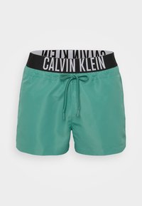 Zelena kupaća šorts s crnim elastičnim pojasom na kojem je logo "Calvin Klein". Uključuje prednju vezicu za prilagodljivost.