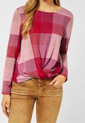Vrouw draagt een roze en rood geruit topje met lange mouwen, gedraaid in de taille, met lichtbruine nauwsluitende broek, hand in de zak.