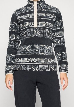 Person trägt einen schwarz-weiß gemusterten Columbia Pullover-Fleece mit Knopfleiste am Kragen und schlichten schwarzen Hosen vor hellem Hintergrund.