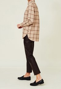 Camisa de cuadros beige de manga larga con una abertura lateral, combinada con pantalones de cuero negros ajustados y mocasines negros. Cuenta con un cuello abotonado.