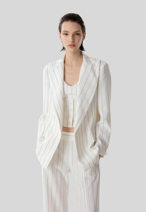 Giovane donna con capelli corti e bagnati che indossa un completo a righe bianche composto da blazer oversize, top corto abbottonato e pantaloni abbinati, in piedi con le mani nelle tasche.