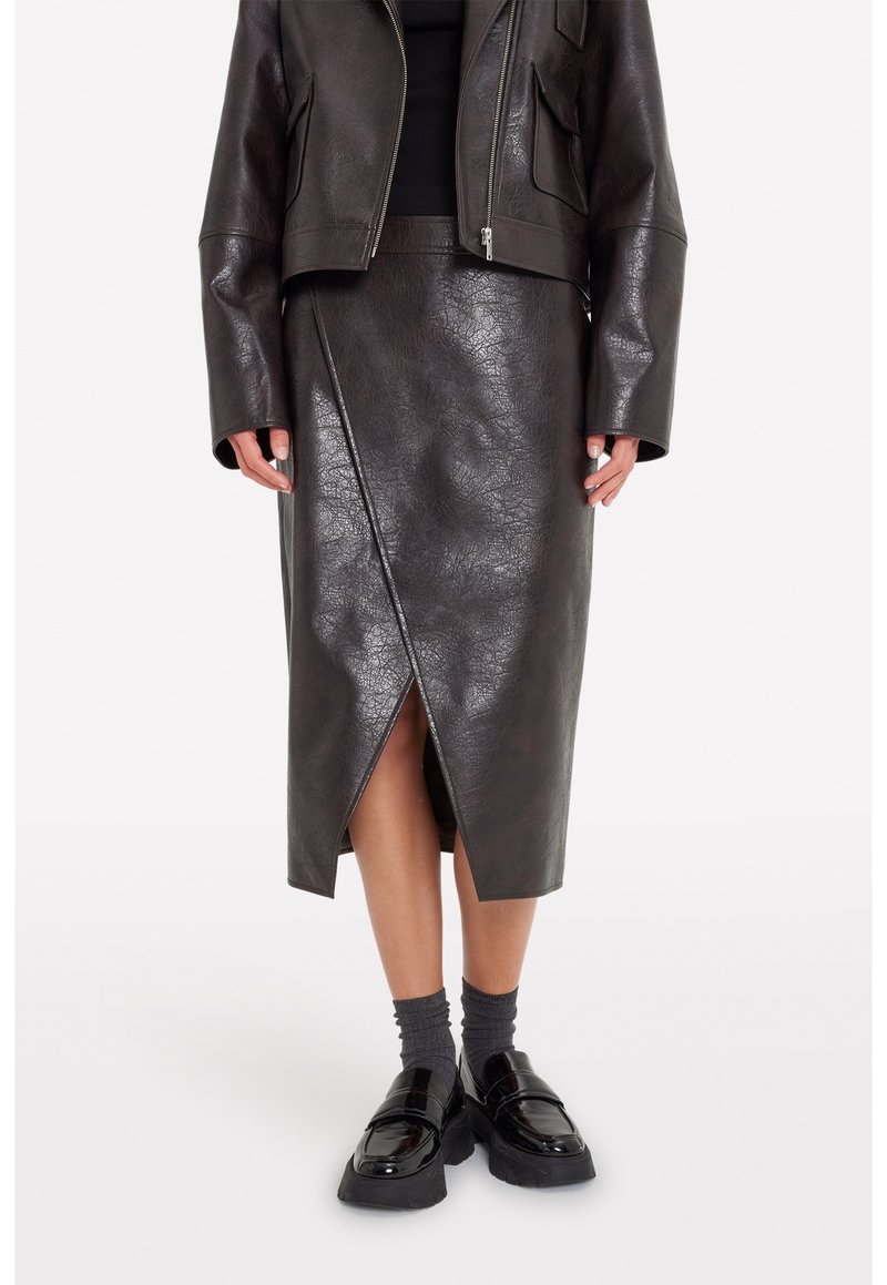 Vue rapprochée d'une personne portant une veste en cuir noire, une jupe en cuir noire drapée avec une fente avant, des chaussettes grises et des mocassins noirs épais.