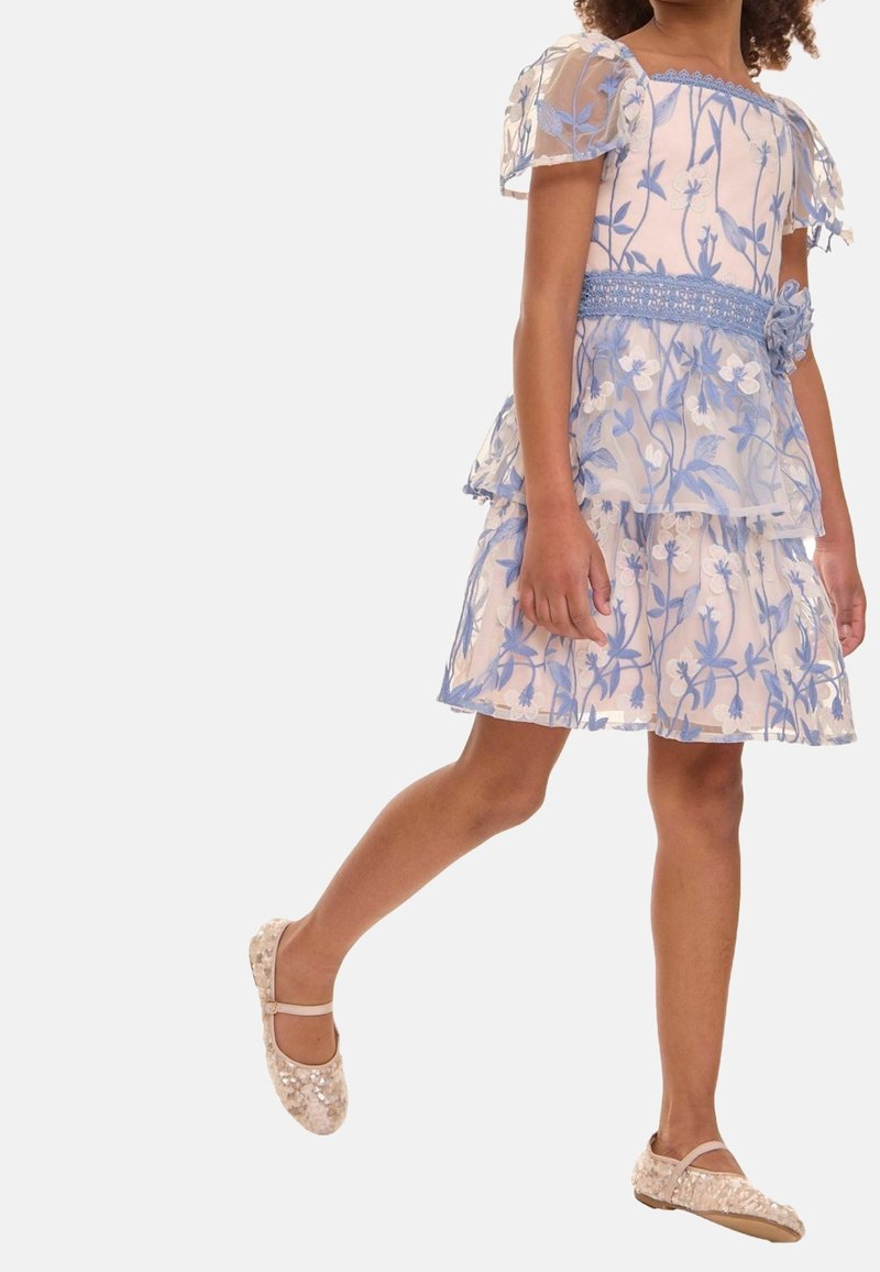 Enfant portant une robe florale lavande avec des manches bouffantes transparentes et des chaussures Mary Jane beige à paillettes, debout avec une jambe tendue.