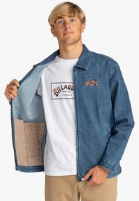 Denim jacket in dark blue with a button closure and two side pockets, lined with a patterned fabric. White t-shirt underneath with logo.