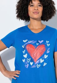 Femme aux cheveux bouclés portant une chemise bleue avec un grand cœur rouge et des cœurs plus petits blancs et violets autour, main sur la hanche.