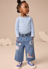 Camicia azzurro chiaro a maniche lunghe con scollo arricciato, culottes in denim con stelle colorate ricamate, una luna e un unicorno. Sneakers blu con dettagli.