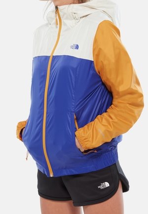 Persoon draagt een blauw, wit en geel North Face jack met capuchon en een zwarte North Face short, handen in de zakken, van opzij bekeken.