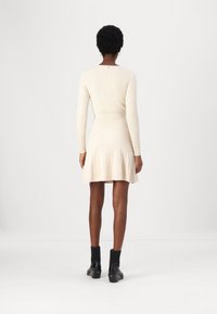 Femme debout de dos portant un haut à manches longues crème côtelé, une jupe plissée assortie et des bottines noires à talons.