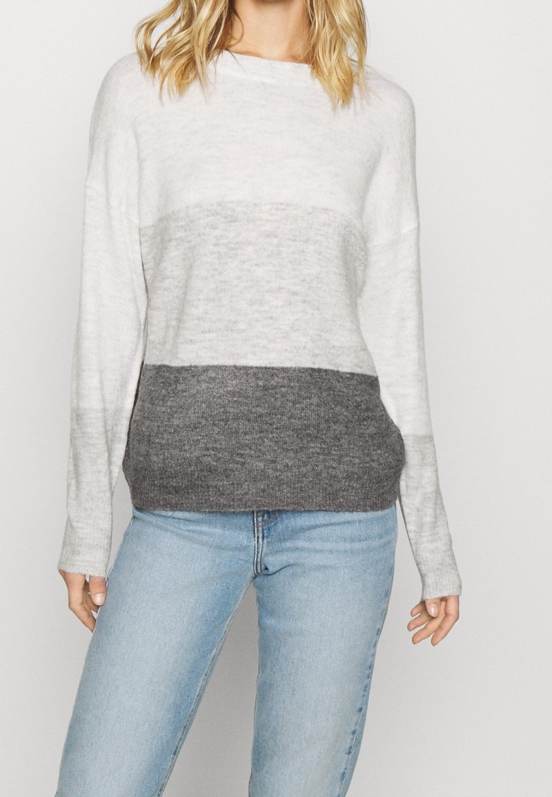Femme portant un pull color-block avec des bandes horizontales blanches, gris clair et gris foncé, associé à un jean bleu clair.