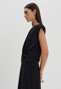 Top nero a maniche corte con dettaglio drappeggiato sul davanti e una texture liscia. Il design ha una vestibilità rilassata e sottili pieghe sui lati.