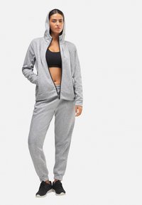 Sudadera gris con cremallera y capucha forrada, combinada con pantalones de chándal a juego. Presenta un ajuste relajado y puños elásticos, llevada con zapatillas negras.