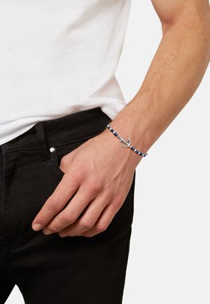 Man met een blauwe kralenarmband met een zilveren ankerbedel, gekleed in een wit shirt en zwarte broek, hand rustend nabij de zak.