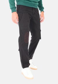 Pantaloni cargo neri in cotone, con molteplici tasche e un dettaglio cucito di rosso sulla gamba sinistra. Abbinati a sneaker bianche.