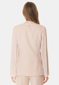 Blazer beige con taglio strutturato e dritto, caratterizzato da maniche lunghe e plissettate e un piccolo dettaglio con logo sul collo sul retro. Tessuto morbido.