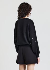 Maglione nero con maniche a sbuffo lunghe, caratterizzato da dettagli in perle bianche lungo i bordi delle maniche. Vestibilità ampia, con orlo a costine. Abbinato a pantaloni corti neri.