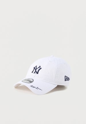 Hvid baseballkasket med navyblåt New York Yankees-logo foran og på siden, buet skygge med signaturdetalje og justerbar rem.