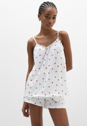 Frau trägt weißes Trägertop und Shorts mit kleinen roten Erdbeermustern, steht vor einem schlichten hellen Hintergrund.
