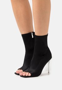 Mustat avonaiset nilkkasukat, joissa on sileä kangaspinta, vetoketju takana ja läpinäkyvä korkea korko. Neliön muotoinen varvasosa.