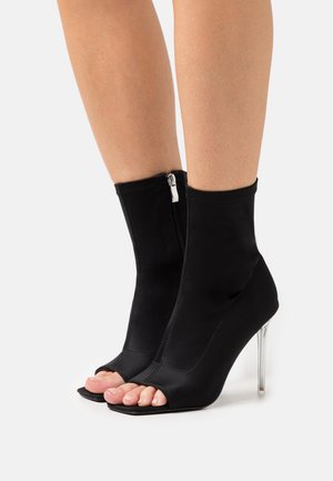 Mustat avonaiset nilkkasukat, joissa on sileä kangaspinta, vetoketju takana ja läpinäkyvä korkea korko. Neliön muotoinen varvasosa.