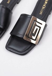 Sandalia deslizante de cuero negro con una tira ancha, que presenta un acento de hebilla de metal dorado de diseño geométrico. Diseño de punta cuadrada con textura suave.