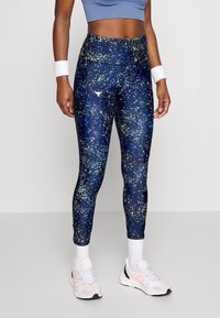 Vrouw in blauw gespikkelde leggings, lichtblauw crop top, witte polsbandjes, witte sokken en witte Adidas sneakers staand tegen een effen achtergrond.
