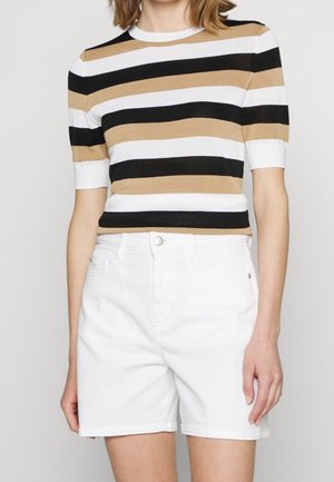 Vrouw in een gestreepte top met korte mouwen in zwart, wit en beige, met hoge witte denim shorts.
