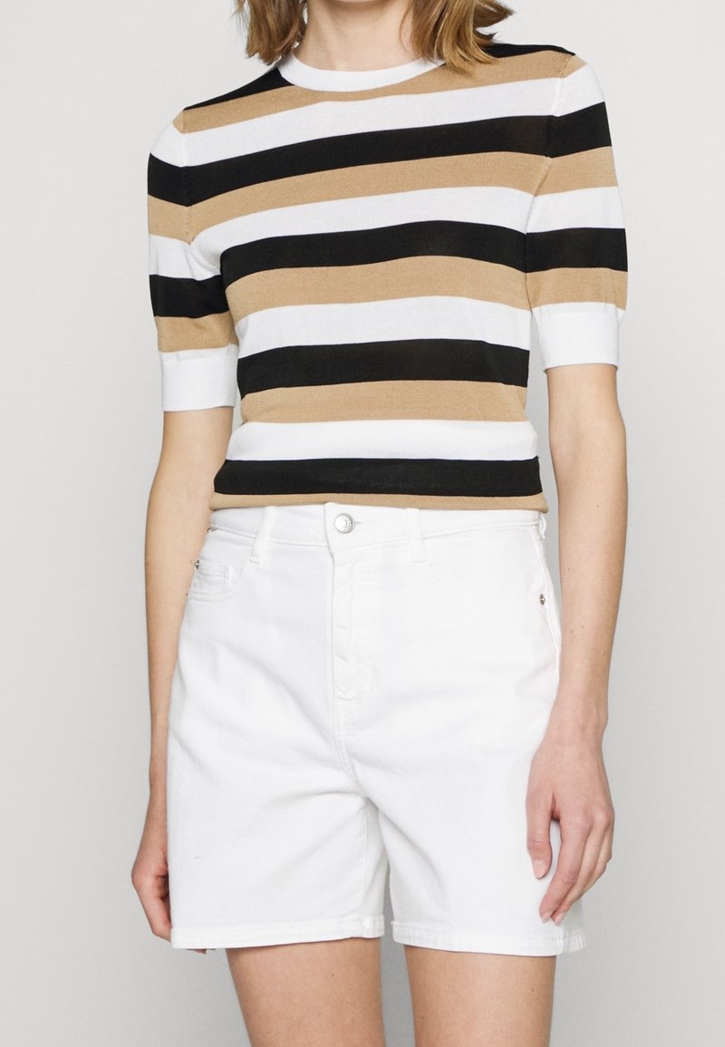 Kvinde iført en kortærmet stribet top i sort, hvid og beige med højtaljede hvide denimshorts.