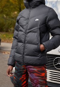 Person som bär en zipad svart Nike-puffjacka och färgglada byxor med mönster i röd, lila och svart, stående bredvid ett vitt fordon.