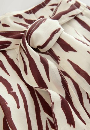 Tkanina z kremowym podkładem, na którym znajdują się burgundowe abstrakcyjne paski. Materiał jest miękki i lekki, z akcentem w postaci zawiązanej kokardy do zapięcia.