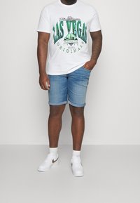 Vitt T-shirt med grön "Las Vegas"-grafik, kombinerad med ljusblå jeansshorts. Bär vita sneakers med gulddetaljer.