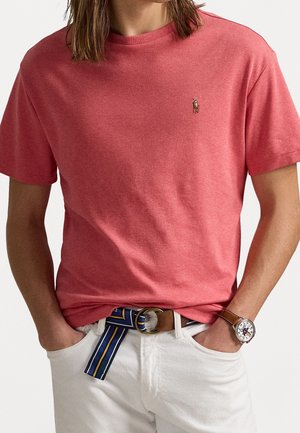 Mann trägt korallenrotes kurzärmliges T-Shirt mit kleinem gesticktem Logo, weiße Hose, braune Lederuhr und blau gestreiften Gürtel, Hände in den Taschen.